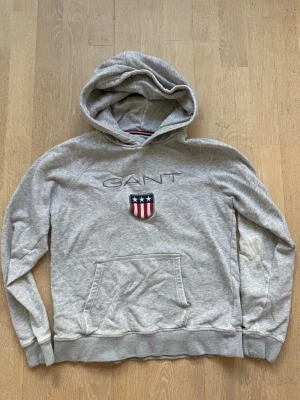 Grå hoodie från GANT med logga - Snygg grå hoodie från GANT med broderad logga och amerikanskt sköldmärke på bröstet. Klassisk känguruficka framtill och huva. Ett hål vid fickan - se bild. 
