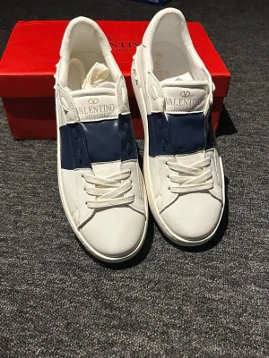 Vita och marinblå sneakers Valentino - Snygga vita sneakers från Valentino med bred marinblå detalj över mitten. Skorna har klassisk låg profil, vita snören och diskret logga på plösen. Tillverkade i slätt läder för en clean och exklusiv look.