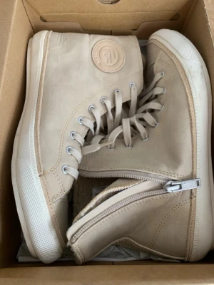 Beige höga sneakers från Lacoste - Snygga beige sneakers i äkta skinn med märket i mocka från Lacoste i högt modell med snörning och dragkedja på sidan. Skorna har en mjuk, ullig insida för extra komfort och en vit sula. Perfekta för dig som vill ha både stil och funktion. Har använt 1 dag bara. 
