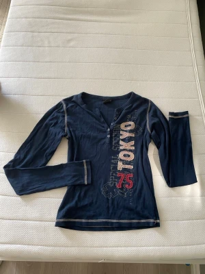 Mörkblå långärmad tröja med tryck - Snygg mörkblå långärmad topp med texten 'TOKYO 75' i vitt och rött framtill. Toppen har knappar vid halsringningen och kontrastsömmar längs ärmar och nederkant. Från Urban outfitters, storlek xs/s