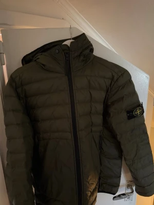 Olivgrön pufferjacka med huva - Säljer en olivgrön pufferjacka från Stone Island med ikonisk patch på vänster ärm. Jackan har huva, dragkedja framtill och quiltad design. Perfekt för kalla dagar och riktigt snygg streetstil. Storlek S/M
