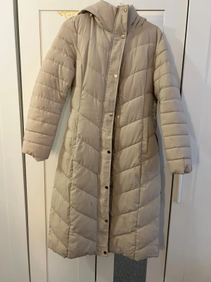 Lång beige dunjacka med huva - Snygg lång beige dunjacka med huva och bälte i midjan. Jackan har quiltat mönster, knappar och dragkedja framtill samt praktiska sidfickor. Perfekt för kalla vinterdagar och ger en stilren look.