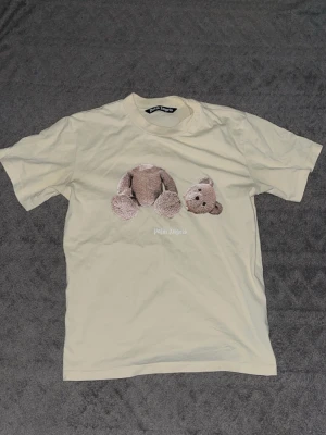 Palm Angels beige t-shirt med björn - Snygg beige t-shirt från Palm Angels med ikoniskt tryck av en nallebjörn med avtagbart huvud på bröstet. T-shirten har rund halsringning och är tillverkad i mjukt bomullsmaterial. Perfekt för dig som gillar streetwear och unika prints.