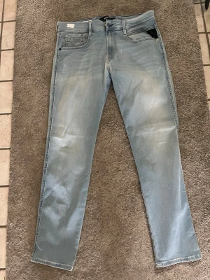 Ljusblå slim fit jeans från Replay - Snygga ljusblå slim fit jeans från Replay med klassisk femficksmodell och diskreta slitningar framtill. Jeansen har en modern tvätt och svarta detaljer på fickan. Perfekta för en trendig och avslappnad stil. Helt nya aldrig använda, nypris 1100 säljs för 499. Storlek: W32 L30