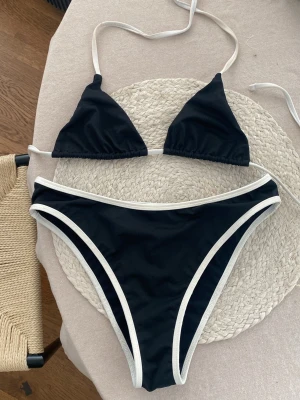 Svart bikini med vita kanter, strl S - Snygg svart bikini med vita kontrastkanter. Överdelen är trekantsmodell med knyt i nacken och ryggen, och underdelen har klassisk passform med vita detaljer längs kanterna. 