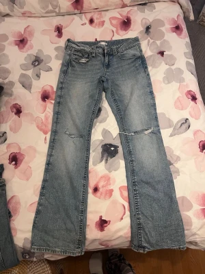 Ljusblå slitna bootcut jeans - Säljer ett par ljusblå bootcut jeans med slitningar och hål vid knäna. Jeansen har klassisk femficksdesign och knappgylf. Perfekta för en avslappnad och trendig look.