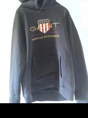 Svart hoodie från Gant med tryck - Svart hoodie från Gant med broderad logga och texten 'American Sportswear' på bröstet. Klassisk huva och stor magficka framtill. Materialet är mjukt och skönt, perfekt för en avslappnad stil. Snygg detalj med röd och vit sköld på bröstet.