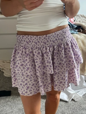 Ljuslila blommig volangkjol - Supersöt kort kjol i ljuslila med små blommor och volangdetaljer. Kjolen har bred resår i midjan och är gjord i ett lätt, luftigt material som passar perfekt till varma dagar. Den är från Gina trixig i storlek m, men passar mig som vanligtvis har storlek s.