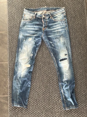 Blå slitna slim jeans från d2 - Säljer ett par blå skinny jeans med slitningar och hål på framsidan. Jeansen har klassisk femficksmodell, låg midja och ljusa tvättade partier för en trendig look. Perfekta för dig som gillar en avslappnad och edgy stil.
