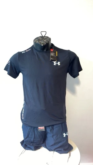 Svart träningsset från Under Armour - Snyggt svart träningsset från Under Armour med t-shirt och shorts. Båda plaggen har logga tryckt i vitt och är tillverkade i ett lätt och snabbtorkande material som passar perfekt för gymmet eller löpning.