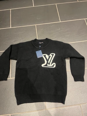 Svart stickad tröja lw - Snygg svart stickad tröja från Lw med stort vitt Lw logo på bröstet. Tröjan har rund halsringning, ribbade muddar och är tillverkad i mjukt material. Perfekt för dig som vill ha en exklusiv och trendig look.