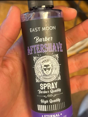 Barber Aftershave Spray från East Moon - Barber Aftershave Spray från East Moon med svart och lila etikett. Flaskan har en cool retrodesign med illustration av en barberare och text i vitt och lila. Sprayen är avsedd för användning efter rakning och ger en fräsch känsla.