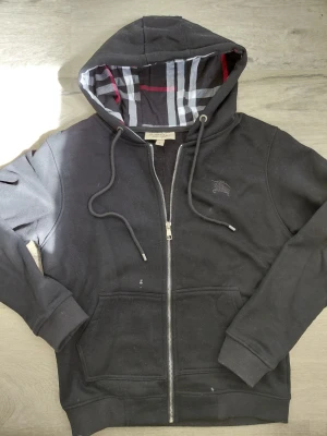 Burberry hoddie  - Snygg svart hoodie med dragkedja och fickor framtill. Huvan har ett klassiskt rutigt mönster i svart, vitt och rött på insidan. Justerbara snören i huvan och broderad logga på bröstet. Perfekt för en avslappnad streetwear-look.