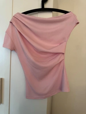 Ljusrosa offshoulder topp - Trendig ljusrosa topp med offshoulder-design och ribbad struktur. Toppen har kort ärm och är tillverkad i ett mjukt, stretchigt material som sitter snyggt på kroppen. Perfekt för dig som vill ha en enkel men stilren look.