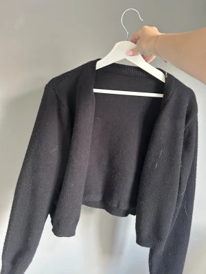 Svart  kofta - En svart, cropped kofta med lång ärm och öppen framtill. Koftan är stickad i ett mjukt material och har ribbade muddar vid ärmslut och nederkant. Knapparna kan du välja själv om du vill visa när du har på dig den. Perfekt att slänga över en topp för en chill look.