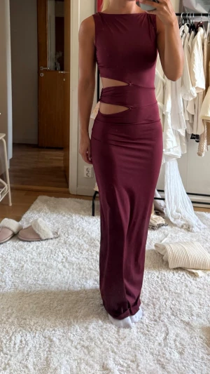 Vinröd långklänning med cut-outs - Vinröd långklänning i figurnära modell med trendiga cut-outs vid midjan. Klänningen är ärmlös och har en rund halsringning. Använd en gång.