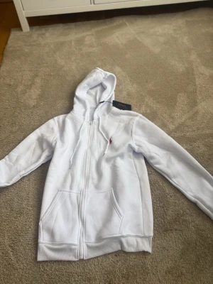Vit hoodie med dragkedja och huva - Säljer en stilren vit hoodie med dragkedja framtill och två stora fickor. Klassisk huva med snören och ribbade muddar vid ärmar och nederkant. Liten broderad logga i rött på bröstet. Perfekt för en clean och avslappnad look.