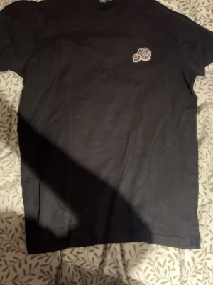 Svart Moncler t-shirt med logga - Snygg svart t-shirt från Moncler med klassisk logga broderad på bröstet. T-shirten har rund hals och korta ärmar, perfekt för en clean och stilren look. Materialet känns mjukt och bekvämt mot huden. Viktigt det står XL men den passar M 
