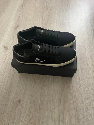 Svarta Saint Laurent sneakers - Svarta sneakers från Saint Laurent med vit broderad logga på sidan och vita sulor. Skorna har klassisk låg profil, svarta skosnören och detaljer i textil och läder. Perfekta för en clean och stilren look.