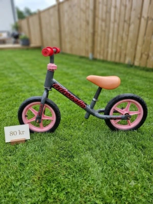 Grå och rosa balanscykel - Säljer en balanscykel i grått med rosa detaljer och orange sadel. Cykeln har två hjul med rosa ekrar och svarta däck, samt röda handtag på styret. Perfekt för att lära sig balansen innan vanlig cykel.