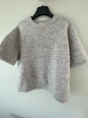 Grå stickad kortärmad tröja - Mysig grå stickad tröja med korta ärmar och rund halsringning. Tröjan har en enkel och avslappnad passform, perfekt att styla med jeans eller kjol. Ribbad kant vid halsen ger en snygg detalj.