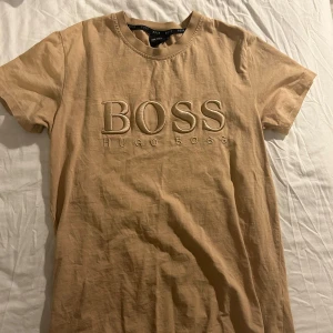 Beige t-shirt från Hugo Boss - Säljer en snygg beige t-shirt från Hugo Boss i bomull. Den har korta ärmar och ett stort BOSS-tryck på framsidan. Perfekt för en avslappnad stil eller att klä upp med en kavaj. Passar både till vardags och fest!
