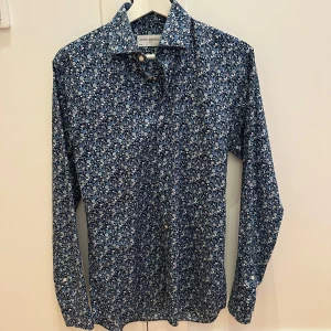 Blommig skjorta från John Henric - Säljer en snygg blommig skjorta från John Henric i storlek 37. Skjortan är blå med ett detaljerat blommönster i vitt och ljusblått. Den har långa ärmar och knappar framtill. Perfekt för både vardag och fest! 🌸