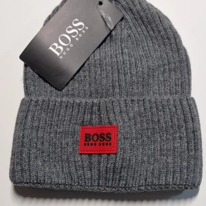 Grå ribbad mössa från Hugo Boss - Snygg och stilren grå ribbad mössa från Hugo Boss. Den har en röd broderad logga framtill som ger en cool kontrast. Perfekt för att hålla värmen under kyliga dagar. Mössan är i nytt skick och passar perfekt till både vardag och mer dressade tillfällen.