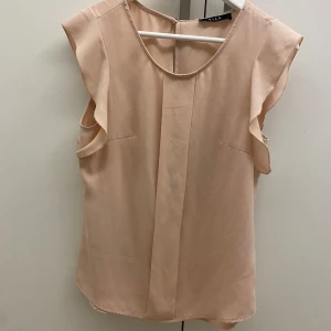 Beige ärmlös topp från Vila - Säljer en elegant beige ärmlös topp från Vila i storlek S. Toppen har en mjuk och luftig design med volangdetaljer vid ärmhålen. Perfekt för både vardag och festliga tillfällen. Den är i mycket bra skick och redo för en ny garderob!