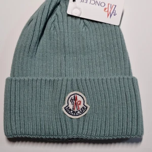 Grön ribbad mössa  - Snygg och stilren grön ribbad mössa från Moncler. Perfekt för kyliga dagar med sitt mjuka och varma material. Den har en uppvikt kant och ett broderat Moncler-märke framtill. Passar både till vardags och mer uppklädda tillfällen.