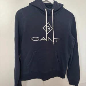 Mörkblå hoodie från GANT - Säljer en snygg mörkblå hoodie från GANT i mycket bra skick. Den har en design med vit logga på framsidan och justerbar huva med snören. Den har inte använts så mycket därav priset, samt inga skador 
