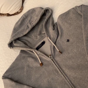 Grå hoodie från Morris - Säljer en snygg och bekväm grå hoodie från Morris. Den har en dragkedja framtill och en liten broderad logga på bröstet. Perfekt för en avslappnad stil och passar till alla årstider. Huvan har snören med snygga detaljer i ändarna. Perfekt för både vardag och chill.