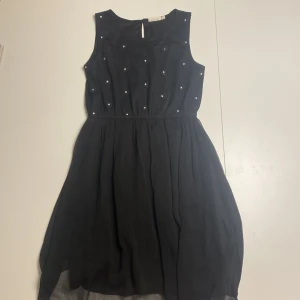 Svart ärmlös klänning från KappAhl - Säljer en elegant svart ärmlös klänning från KappAhl i storlek 158/164. Klänningen har fina blommiga detaljer med små pärlor i mitten, vilket ger en extra touch av glamour. Perfekt för festliga tillfällen! 🖤