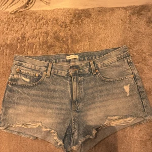 Blå lågmidjade jeansshorts från Gina  - Säljer ett par snygga blå lågmidjade  jeansshorts från Gina tricot i storlek 42. De har en  slitning och fransiga kanter för en avslappnad look. Perfekta för sommardagar eller festivaler💕