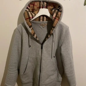 Grå Burberry hoodie   - Säljer en snygg grå hoodie från Burberry i storlek L. Den har en klassisk dragkedja och en huva med det ikoniska Burberry-mönstret på insidan. Perfekt för både vardag och chill. Den är i mycket bra skick och superbekväm att ha på sig. Passar perfekt till höst och vår!