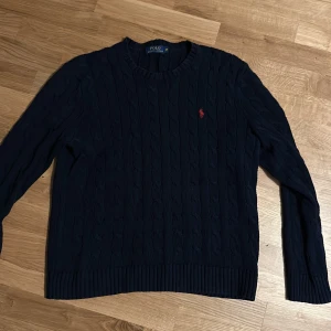 Mörkblå stickad tröja från Ralph Lauren - Säljer en snygg mörkblå stickad tröja från Ralph Lauren. Den har ett klassiskt kabelstickat mönster och en liten röd logga på bröstet. Perfekt för höst och vinter. Skicket är helt okej eftersom den använts länge skulle säga ungefär 8/10 men den sitter fortfarande fint. Bara att skriva vid funderingar är också öppen till byten. (Inga returer)