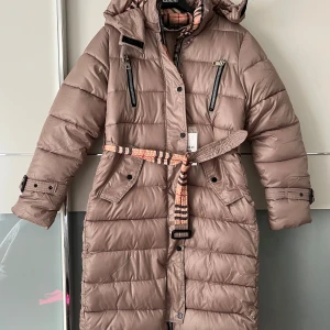 Beige dunjacka med bälte - Säljer en superfin beige dunjacka från Burberry. Den är lång och har ett snyggt bälte i midjan för en figursydd look. Jackan har en huva och dragkedja framtill, samt praktiska fickor med knappar. Perfekt för kalla vinterdagar! 🧥❄️