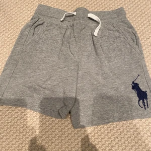 Grå shorts från Polo Ralph Lauren - Säljer ett par snygga grå shorts från Polo Ralph Lauren i storlek 5 år. Detaljerad med den ikoniska Polo-loggan i mörkblått på benet. Perfekta för en avslappnad stil!