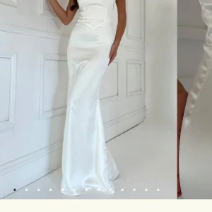Vit lång balklänning i satin - Elegant vit långklänning i satin med en tight passform. Klänningen har en vacker knapprad i ryggen och är axelbandslös, vilket ger en stilren look. Perfekt för bal och materialet ger en lyxig känsla och klänningen har en tidlös design. Dock finns det lite fläckar och slitningar längst ner men det går att sy upp då den är väldigt låg och tvätta bort😊