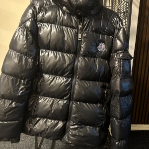 Svart pufferjacka från Moncler - Säljer en snygg svart pufferjacka från Moncler i mycket bra skick. Jackan är perfekt för vintern med sitt quiltade mönster och långa ärmar. Den har en praktisk dragkedja och en huva för extra värme. Passar både till vardags och festliga tillfällen. 🖤