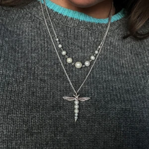 Maria Nilsdotter Silverhalsband med pärlor och trollslända - Säljer ett stilrent silverhalsband med en unik trollsländeberlock och vackra pärlor. Perfekt för att ge en elegant touch till din outfit. Kedjan är i silver och berlocken har en rad av små pärlor. Passar både till vardag och fest! Väldigt bra skick