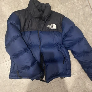 Blå och svart pufferjacka från The North Face - Säljer en snygg och varm pufferjacka från The North Face i blått och svart. Jackan har en hög krage och dragkedja framtill, perfekt för kalla vinterdagar. Den är i bra skick. Priset kan även diskuteras 💕