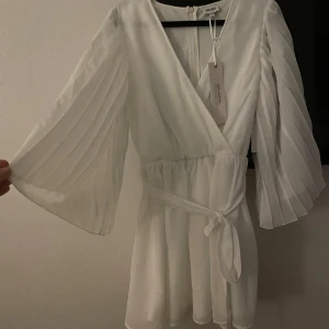 Student Klänning från Bubbleroom - Säljer en superfin vit klänning från Bubbleroom i storlek 38. Den har en omlott-design med ett knytband i midjan och vida, plisserade ärmar. Perfekt för student eller en elegant middag. Klänningen är i nytt skick med taggar kvar. Hör av er för fler bilder eller frågor 🤍