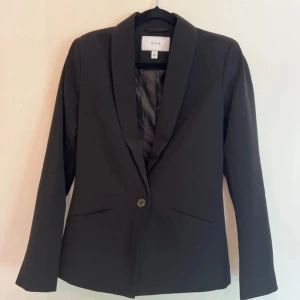 Svart kavaj från Vila - Snygg svart kavaj från Vila i storlek 34. Perfekt för både vardag och fest! Den har en enkel knapp framtill och två fickor. Kavajen är i mycket bra skick och ger en stilren look. Passar perfekt till alla tillfällen. Den är använd en gång