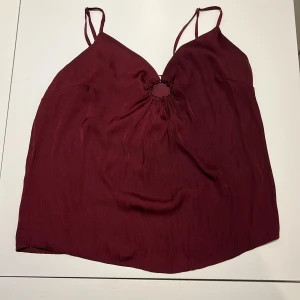 Vinröd linnetopp från About You - Säljer en snygg vinröd linnetopp från About You. Toppen har tunna axelband och en fin detalj med en ring framtill som ger en unik look. Perfekt för sommardagar eller en kväll ute. Materialet känns mjukt och bekvämt mot huden. Står inte storlek men passar mig som är xs/s💞