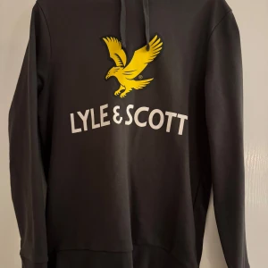 Svart hoodie från Lyle & Scott - Säljer en mörk grå hoodie från Lyle & Scott med deras ikoniska gula logotyp på framsidan. Tröjan har långa ärmar och en bekväm huva. Perfekt för en avslappnad stil.