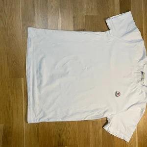 Vit t-shirt från Moncler - Säljer en stilren vit t-shirt från Moncler med ett litet broderat emblem på bröstet. Perfekt för en avslappnad och snygg look. T-shirten är kortärmad och gjord i mjukt bomullsmaterial.