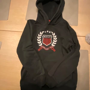 Svart hoodie från GANT - Säljer en svart hoodie från GANT med ett stort tryck på framsidan. Storlek 146/152. Kan fraktas eller hämtas i Skövde mvh Lukas.