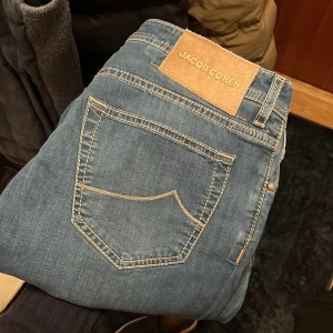 Jacob Cohen jeans  - Snygga blå jeans från Jacob Cohen med klassisk femficksdesign och knappgylf. Jeansen har en stilren look med kontrasterande sömmar och en liten broderad detalj på myntfickan. Perfekta för en avslappnad men stilren outfit.
