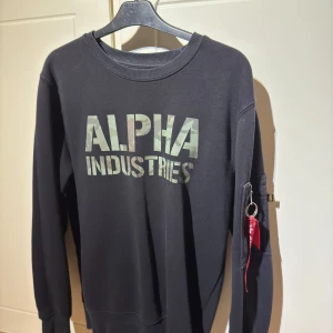 Svart tröja från Alpha Industries - Säljer en svart tröja från Alpha Industries med deras logga i grått på framsidan. Tröjan har långa ärmar och en praktisk ficka med dragkedja på ärmen. Storleken är i S och är i bra skick.